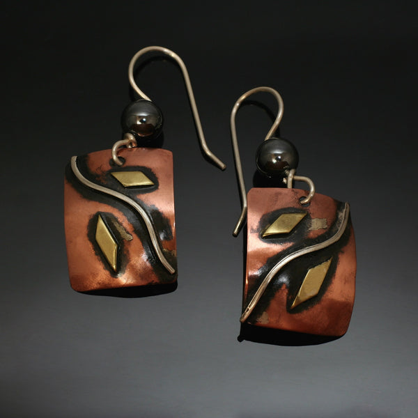 silver wave from the top corner to the opposite bottom corner of copper earrings with black patina and hematite bead