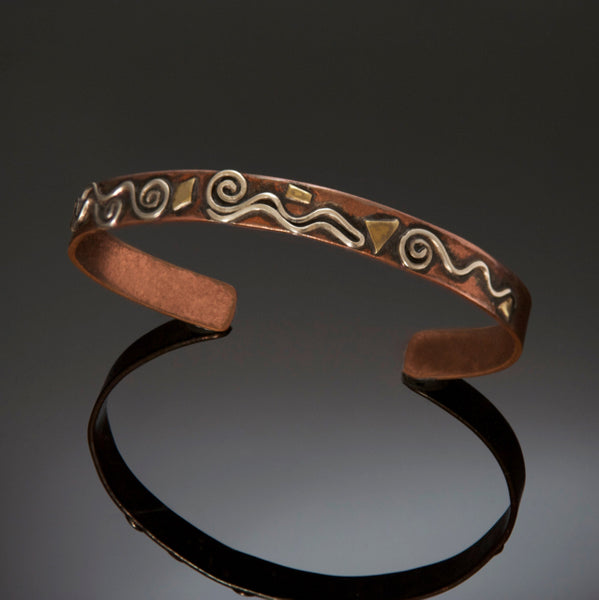 silver spirals on top of a narrow copper bracelet with little brass accents and black patina