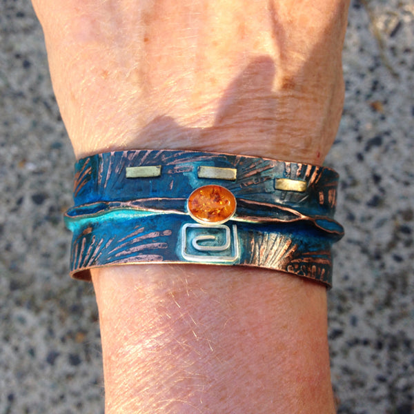 wide folded copper bracelet with embossed fern pattern, square silver and brass with blue-green patina and oval amber.