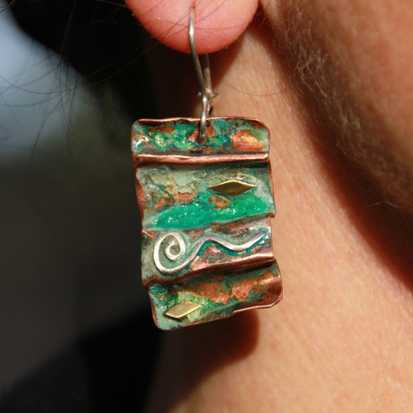 folded copper earrings with silver spiral wave, brass accents and red green patina hanging from an earlobe