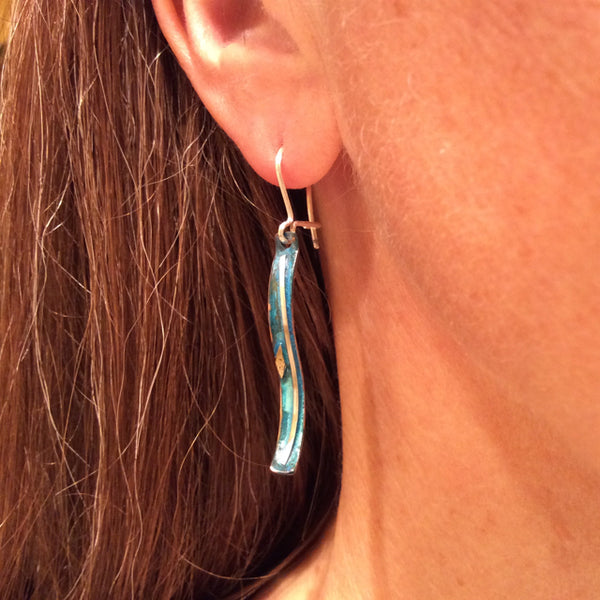 long skinny copper earrings with blue patina, a silver wire down the centre the earring is s-shaped hanging from an earlobe