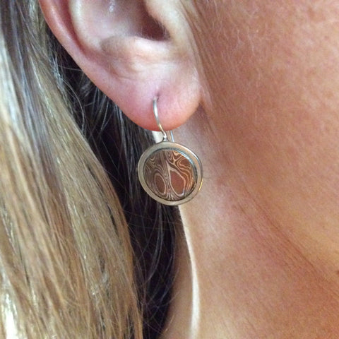 Round silver earrings with inlayed disc of mokume gane on a persons earlobe. Sterling silver hook is soldered onto the earring. The mokume has a wood grain pattern with a dark brown copper patina
