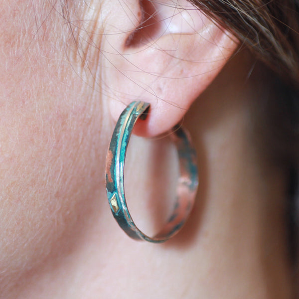 narrow copper hoops with a line of silver wire, brass accents and blue/green patina hanging from an earlobe