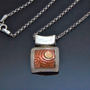 square silver pendant with inlayed copper embossed with spirals and a circle of gold fill framed by silver and japanese style bale.