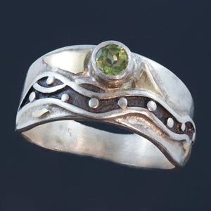 silver ring with faceted peridot surrounded by gold accents and recessed parts oxidized black.