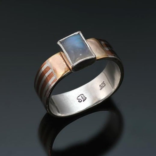 Narrow silver band with silver and copper stripes with a rectangular rainbow moonstone and gold on either side of the stone