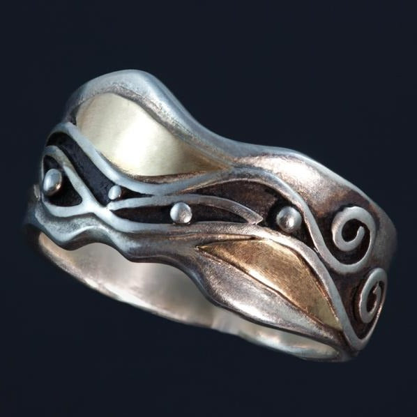 Wide silver ring with uneven edges, waves and spirals on the side, gold accents around the pattern.