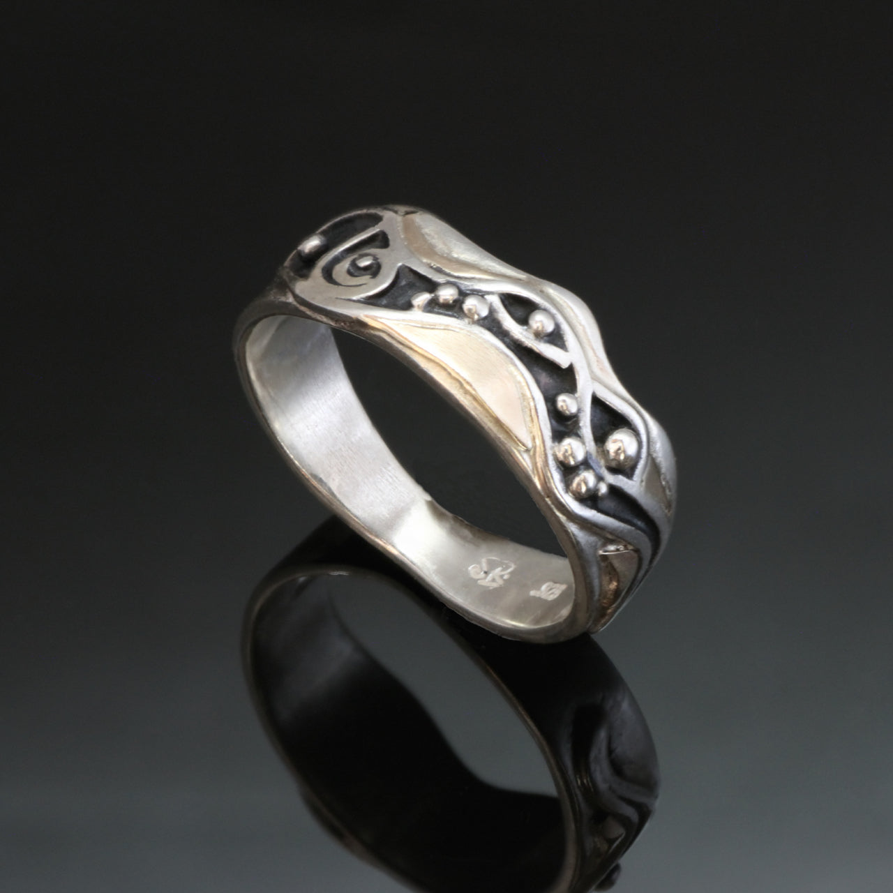 Silver ring with uneven edges, waves, spiral and dots showing a lively water scene. The oxidized pattern has gold accents around it.